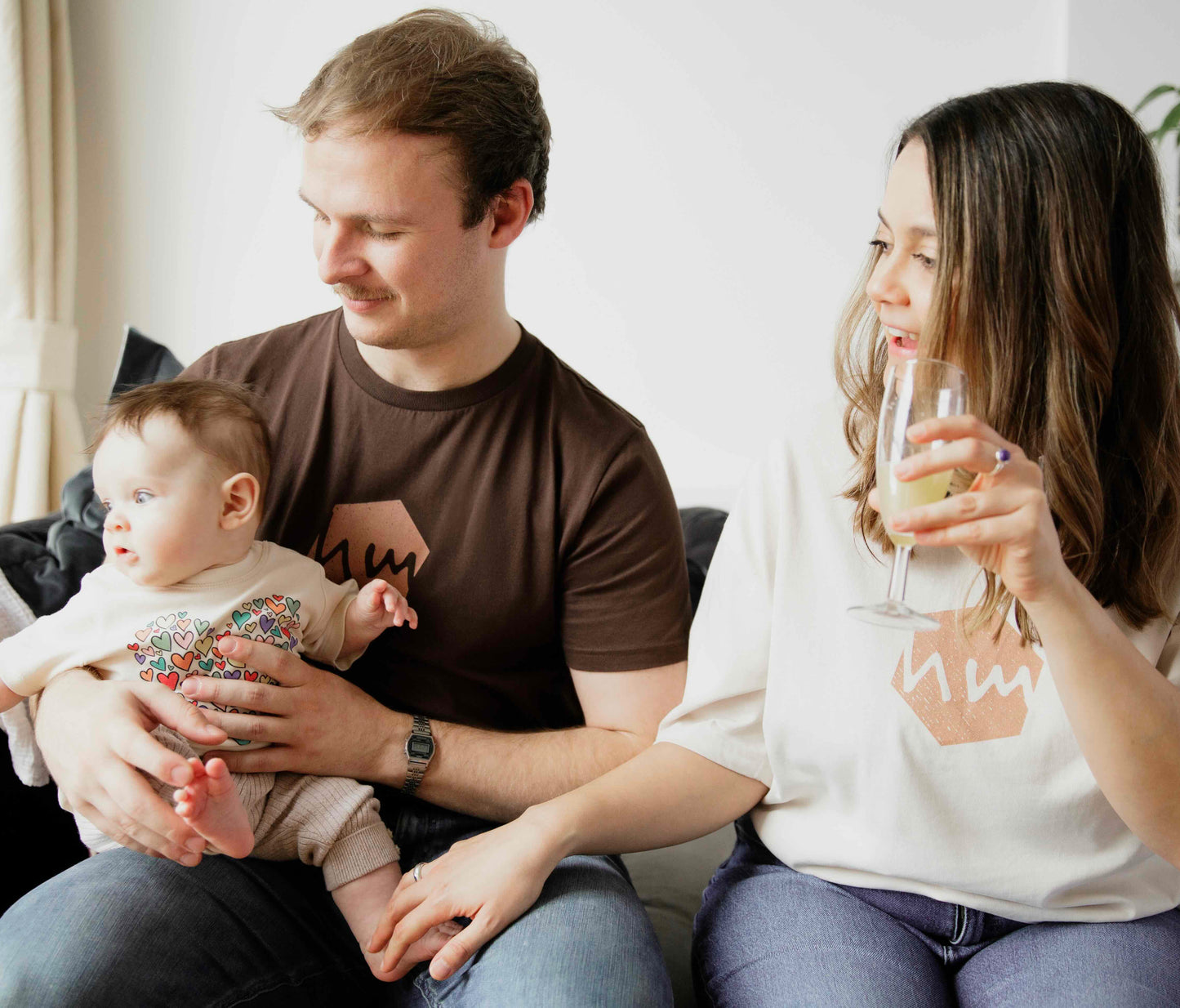 Mother, father and baby wearing human milk t-shirts sitting on a couch, woman holding a glass, with the man holding the baby.