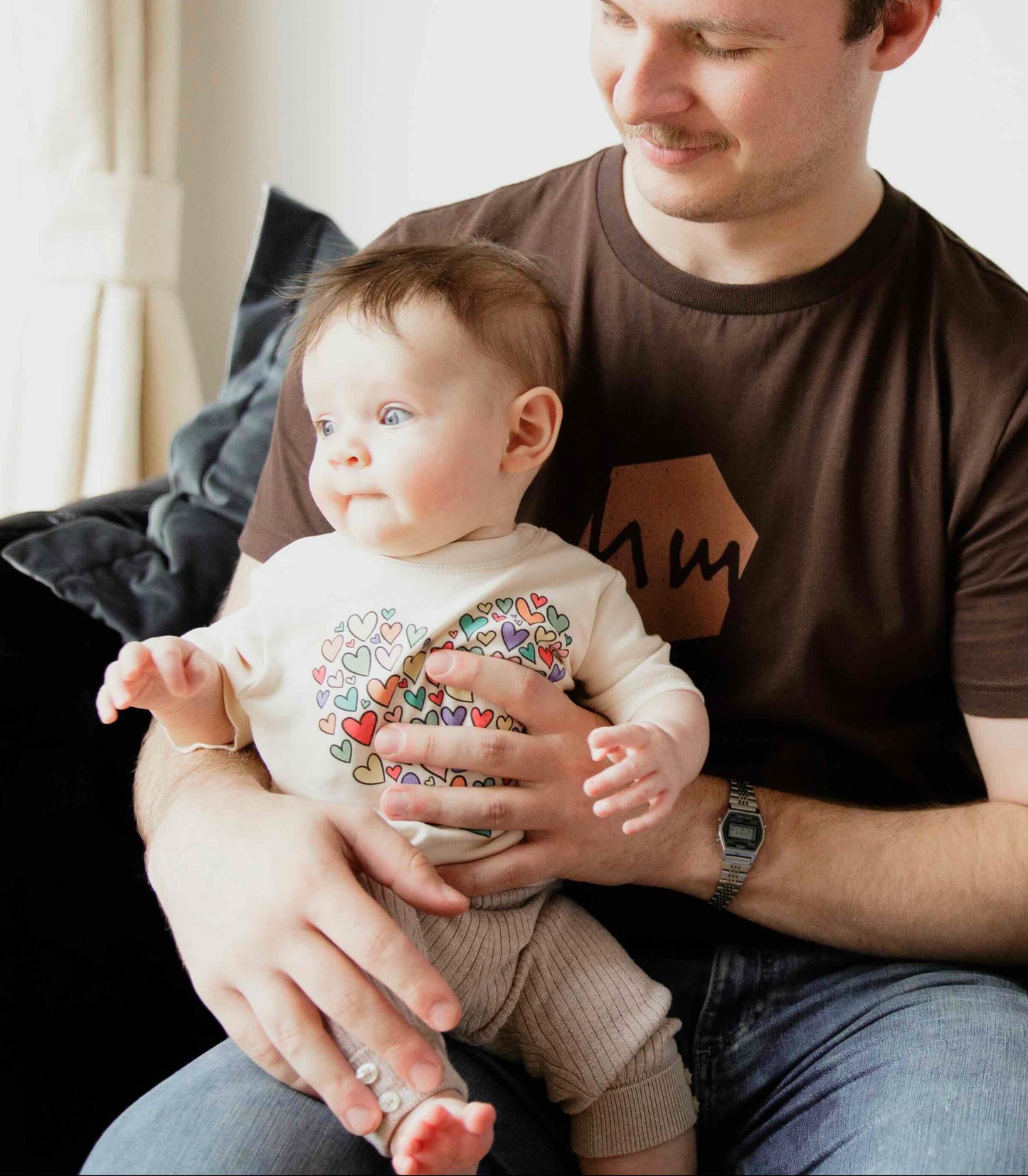 A father sitting on a couch holding his baby. The father is wearing a Human Milk logo T-shirt and the baby is wearing a Human Milk bodysuit
