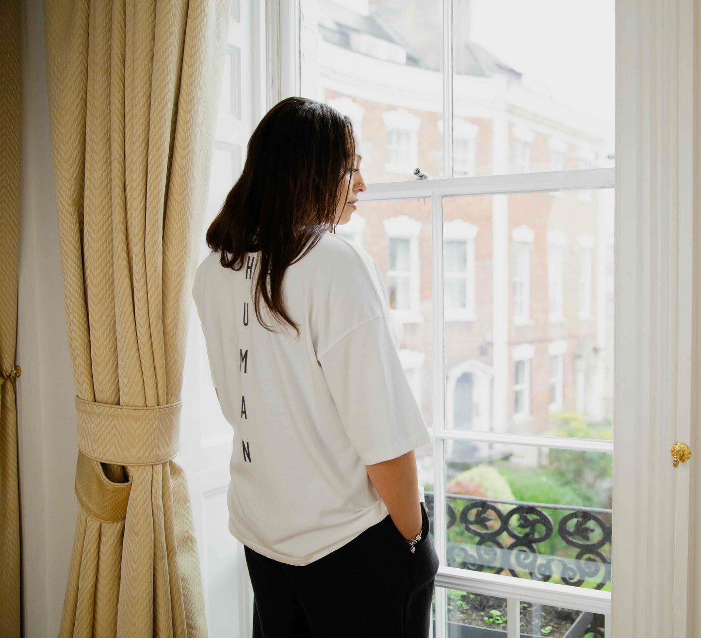 Woman wearing a white human-milk t-shirt with HUMAN text, standing by a window with beige curtains.