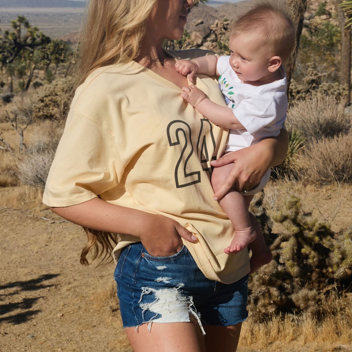 Woman holding a baby in a desert landscape with Joshua Tree in the background, wearing a Human Milk 247 T-shirt.