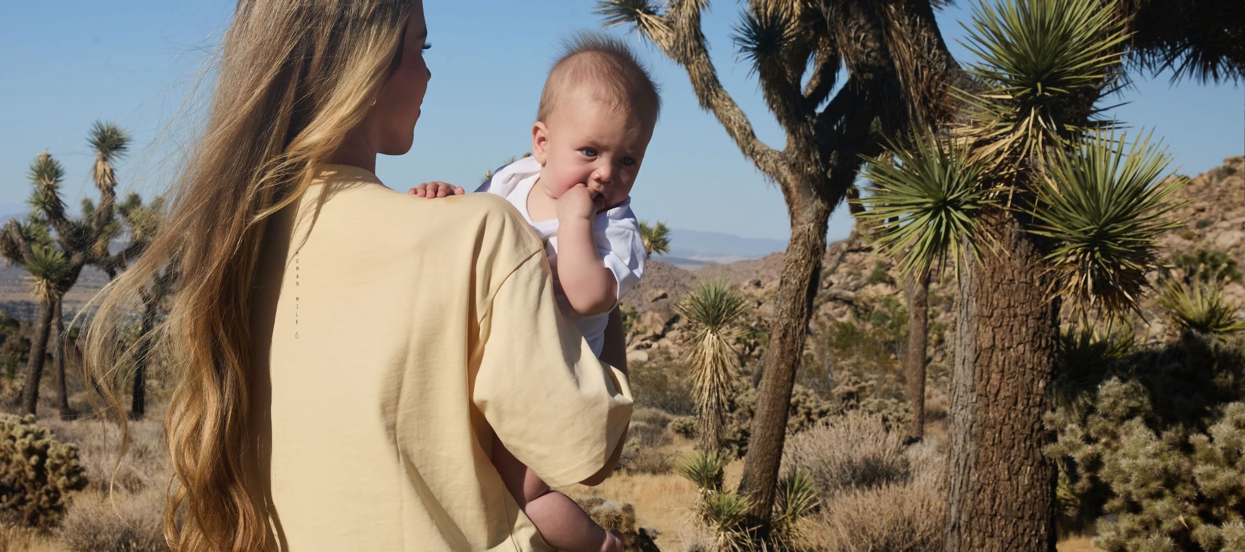 Mother holding her baby in a desert landscape with Joshua trees, wearing a Human Milk T-shirt.
