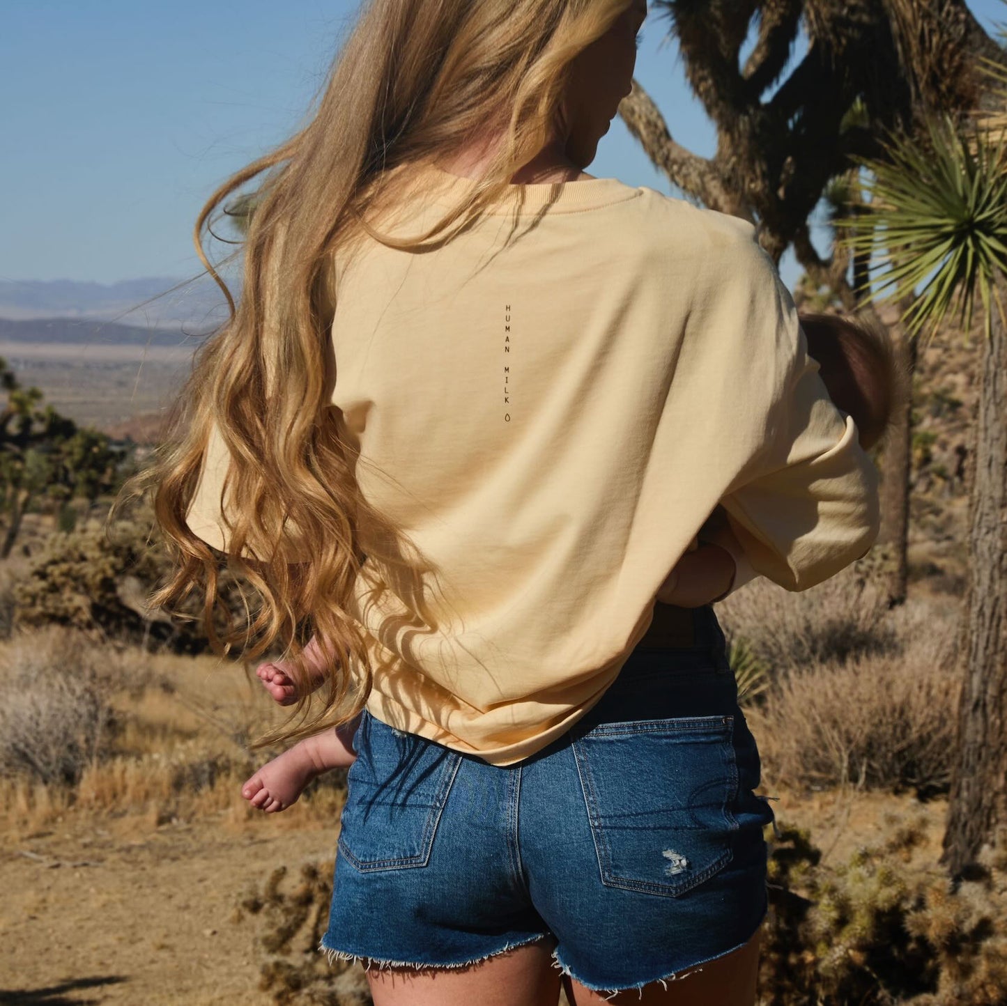 Woman in a desert landscape with Joshua trees, wearing a Human Milk 247 T-shirt, viewed from the back, carrying a baby.