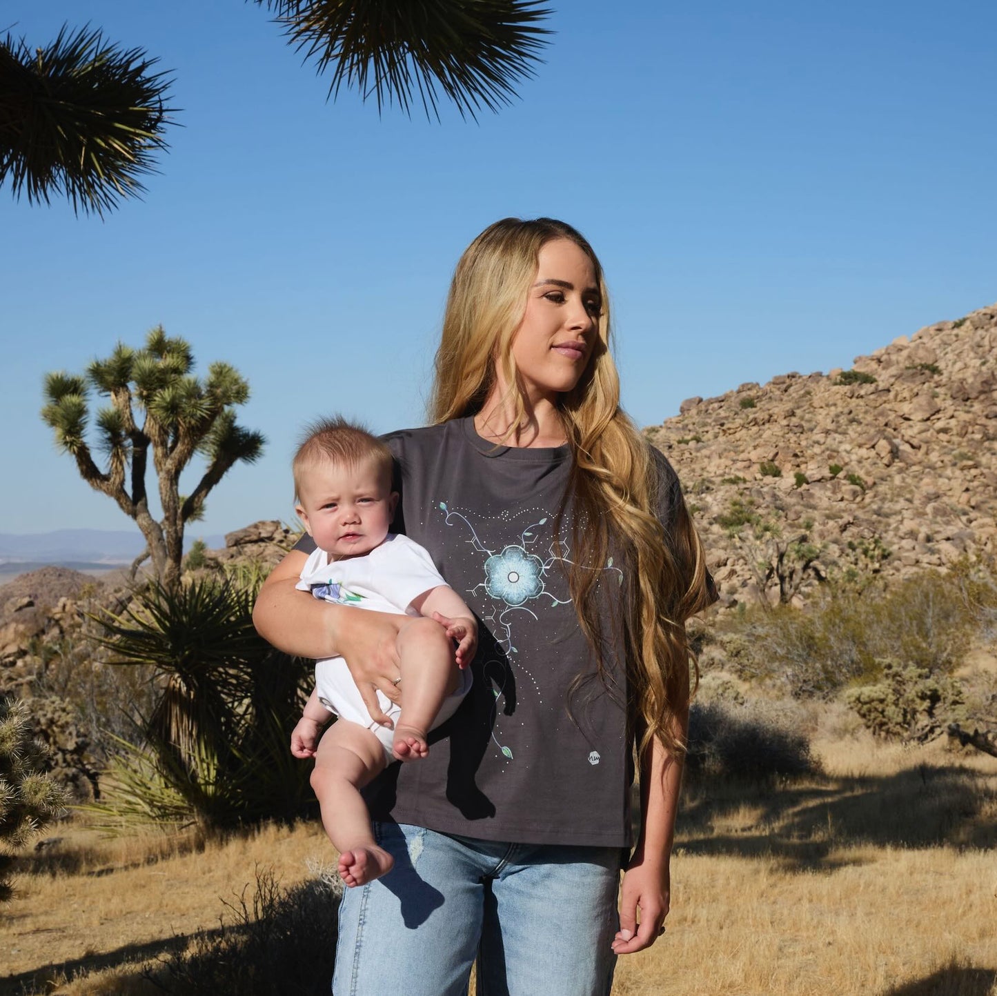 Woman wearing a Human Milk Oxyflower T-shirt holding her baby in a desert landscape with Joshua trees.