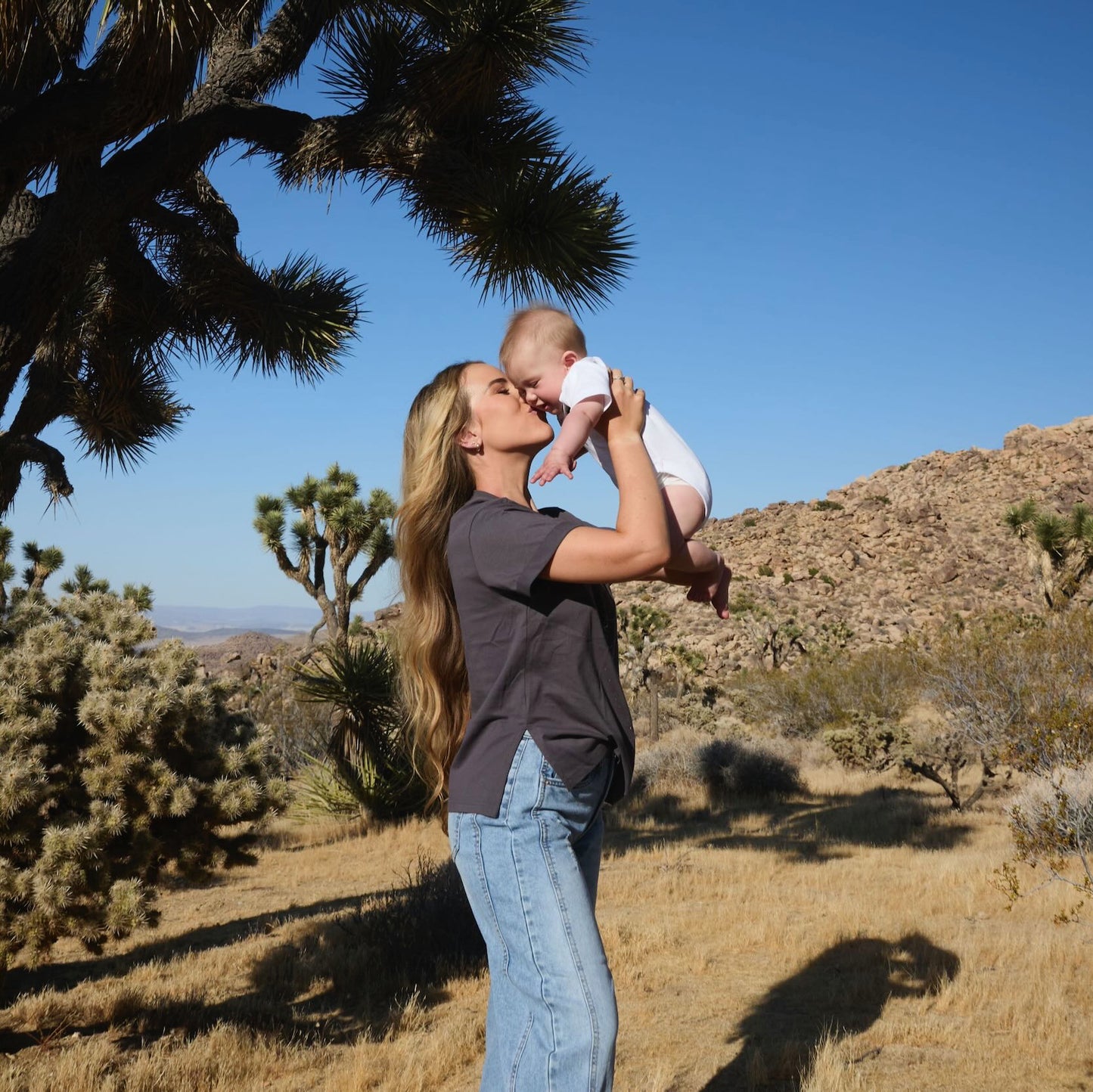 Woman wearing a Human Milk Oxyflower T-shirt, holding her baby in a desert landscape with Joshua trees.