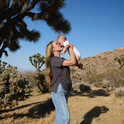 Woman wearing a Human Milk Oxyflower T-shirt, holding her baby in a desert landscape with Joshua trees.