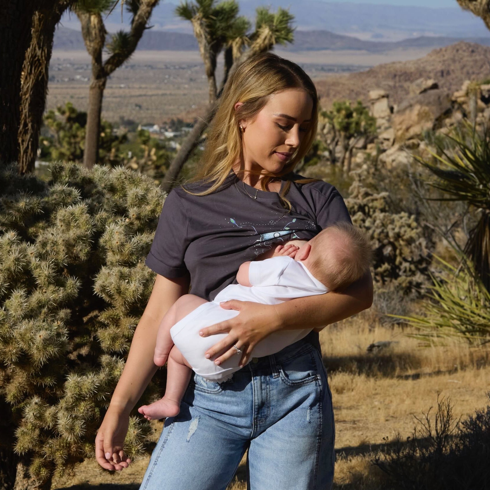 Woman wearing a Human Milk Oxyflower T-shirt, breastfeeding her baby in a desert landscape with Joshua trees.