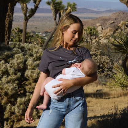 Woman wearing a Human Milk Oxyflower T-shirt, breastfeeding her baby in a desert landscape with Joshua trees.
