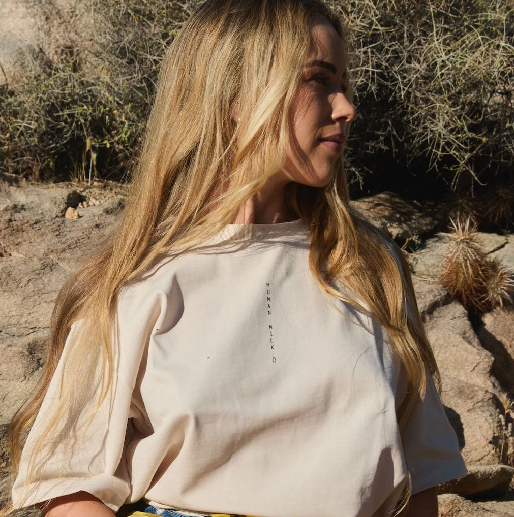 Woman sitting on rocks in a desert-like environment, wearing a Human Milk T-shirt and scarf