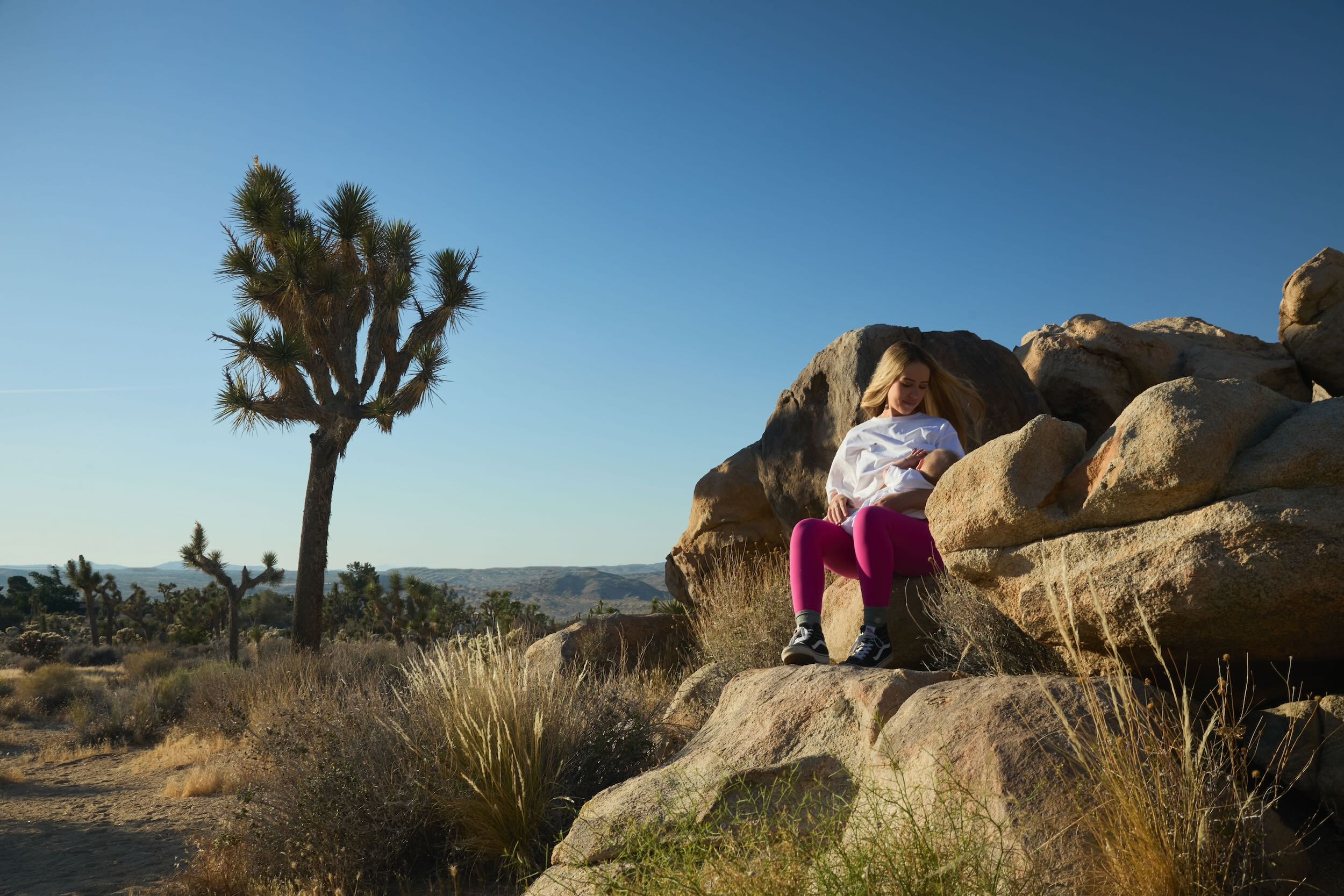 Mother sitting on a rock in a desert landscape with Joshua trees breast-feeding her baby, wearing a Human Milk T-shirt