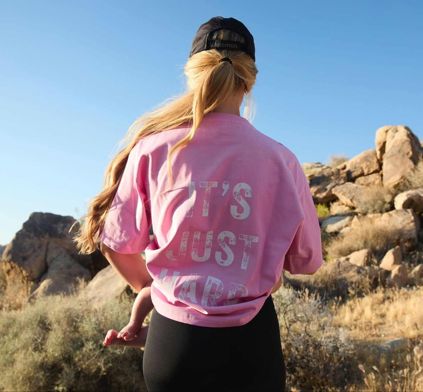 A woman wearing a pink T-shirt with text saying "It's Just Hard", holding a baby, in a desert landscape