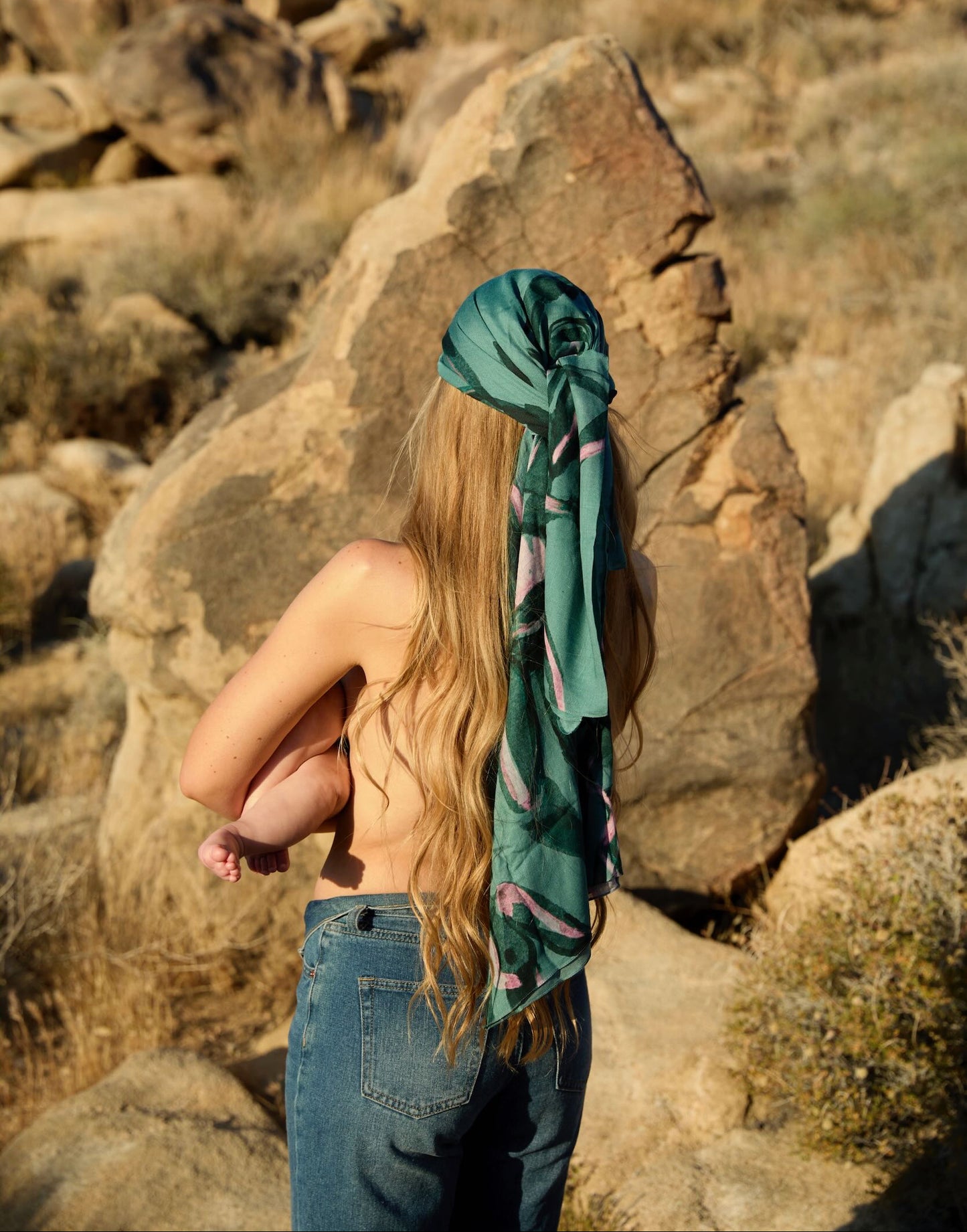 Woman with a green Human Milk x Chloe Trayhurn headscarf standing in a rocky desert landscape, holding her baby, seen from the back