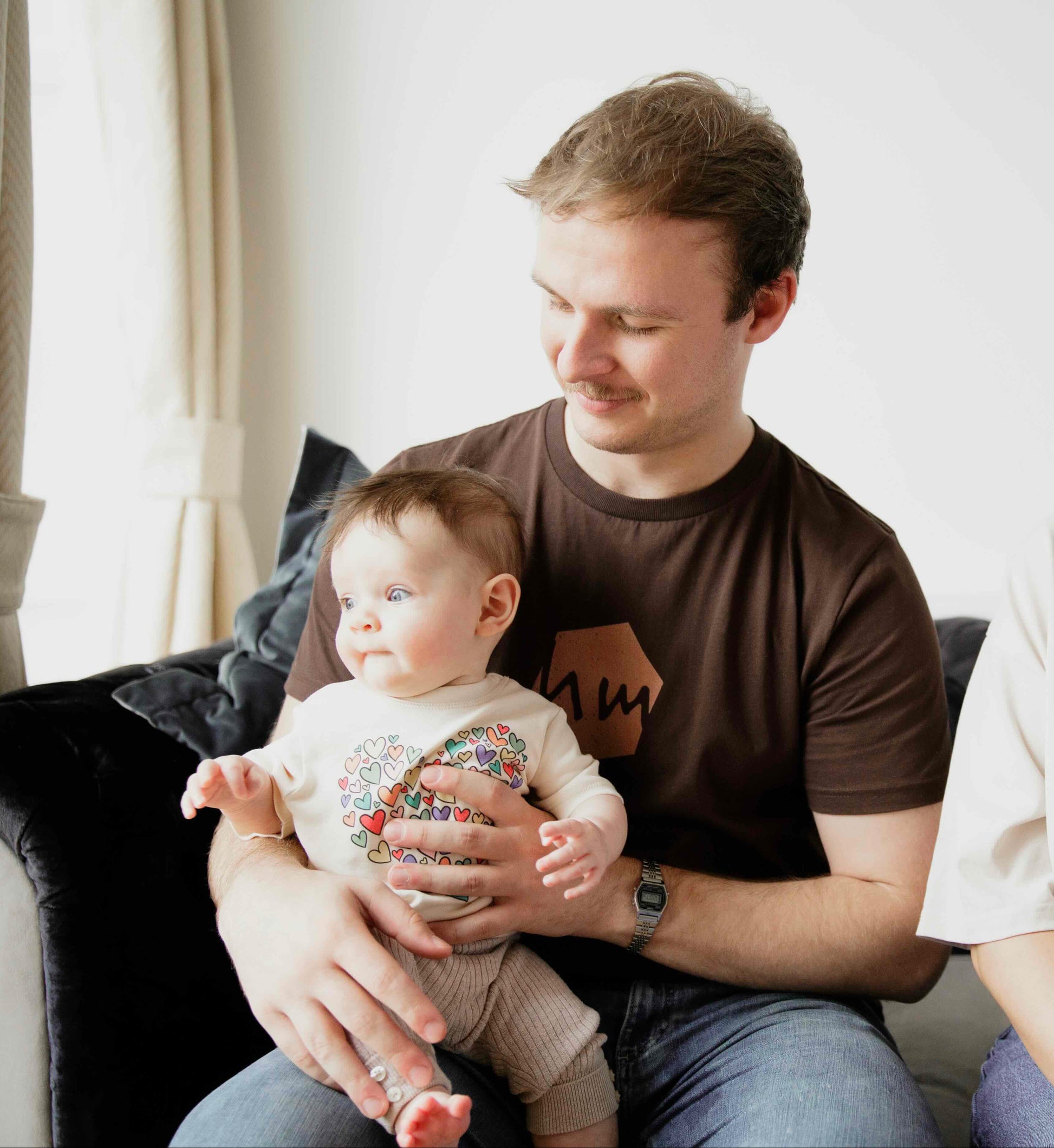 Family of three sitting on a couch with a baby wearing a Human Milk T-shirt held by the father and a woman sitting next to him.