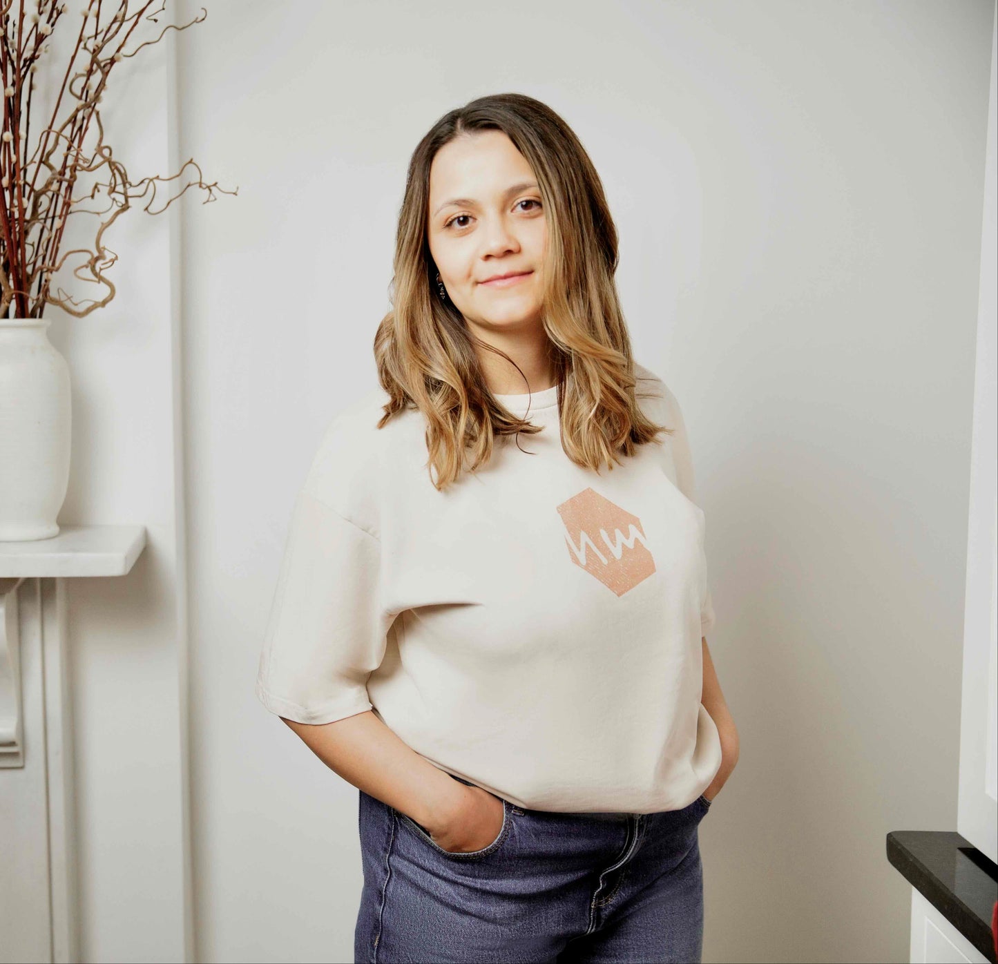 Woman wearing an oat coloured human milk logo t-shirt, standing in a room with a fireplace and decorative vases.
