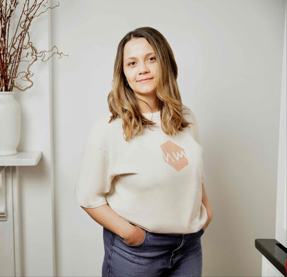 Woman wearing an oat coloured human milk logo t-shirt, standing in a room with a fireplace and decorative vases.