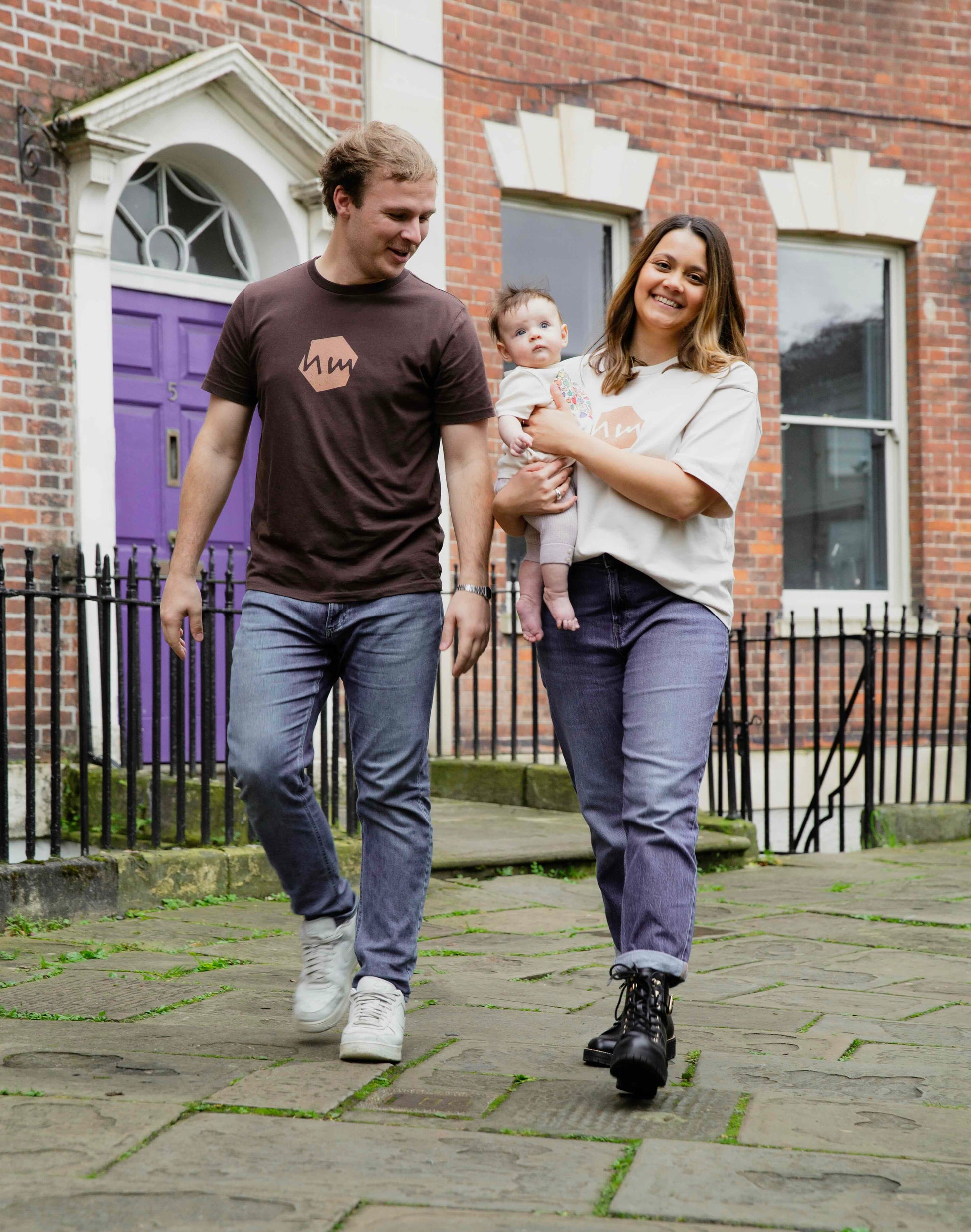 Mother and father walking outdoors in front of a brick building, holding their baby and wearing human milk logo t-shirts.