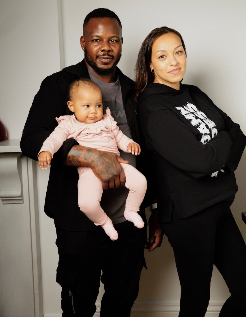 A father holding his baby and his wife leaning into him, smiling. Both parents are wearing Human Milk tops.