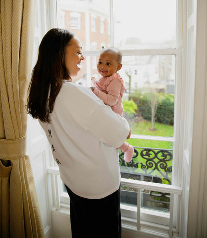 Woman wearing a human milk t-shirt holding her baby by a large window with a view of a garden.