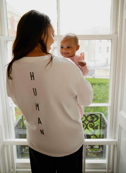 Woman wearing a human milk t-shirt holding a baby in front of a window with 'HUMAN' text on her shirt.