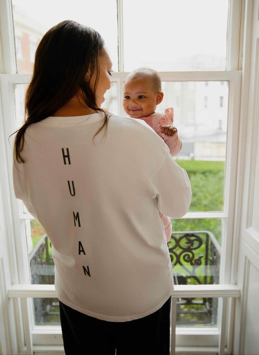 Woman wearing a human milk t-shirt holding a baby in front of a window with 'HUMAN' text on her shirt.