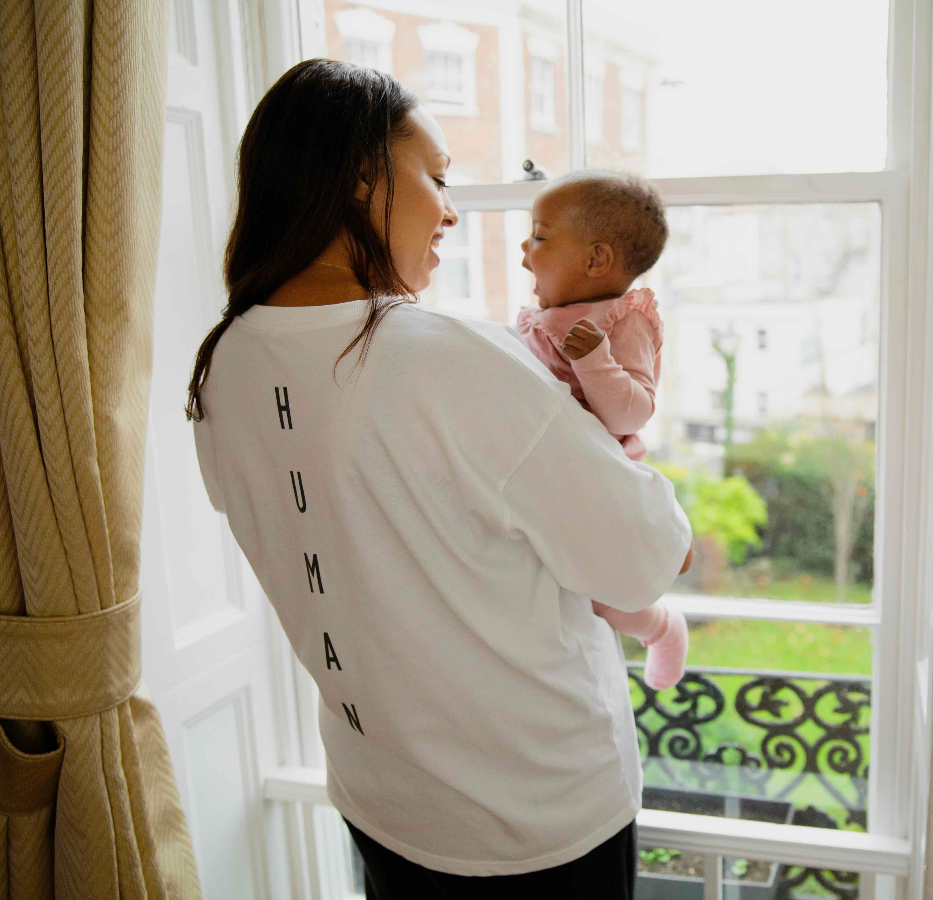 Woman wearing a human milk breastfeeding friendly t-shirt holding her baby by a window with a view of greenery