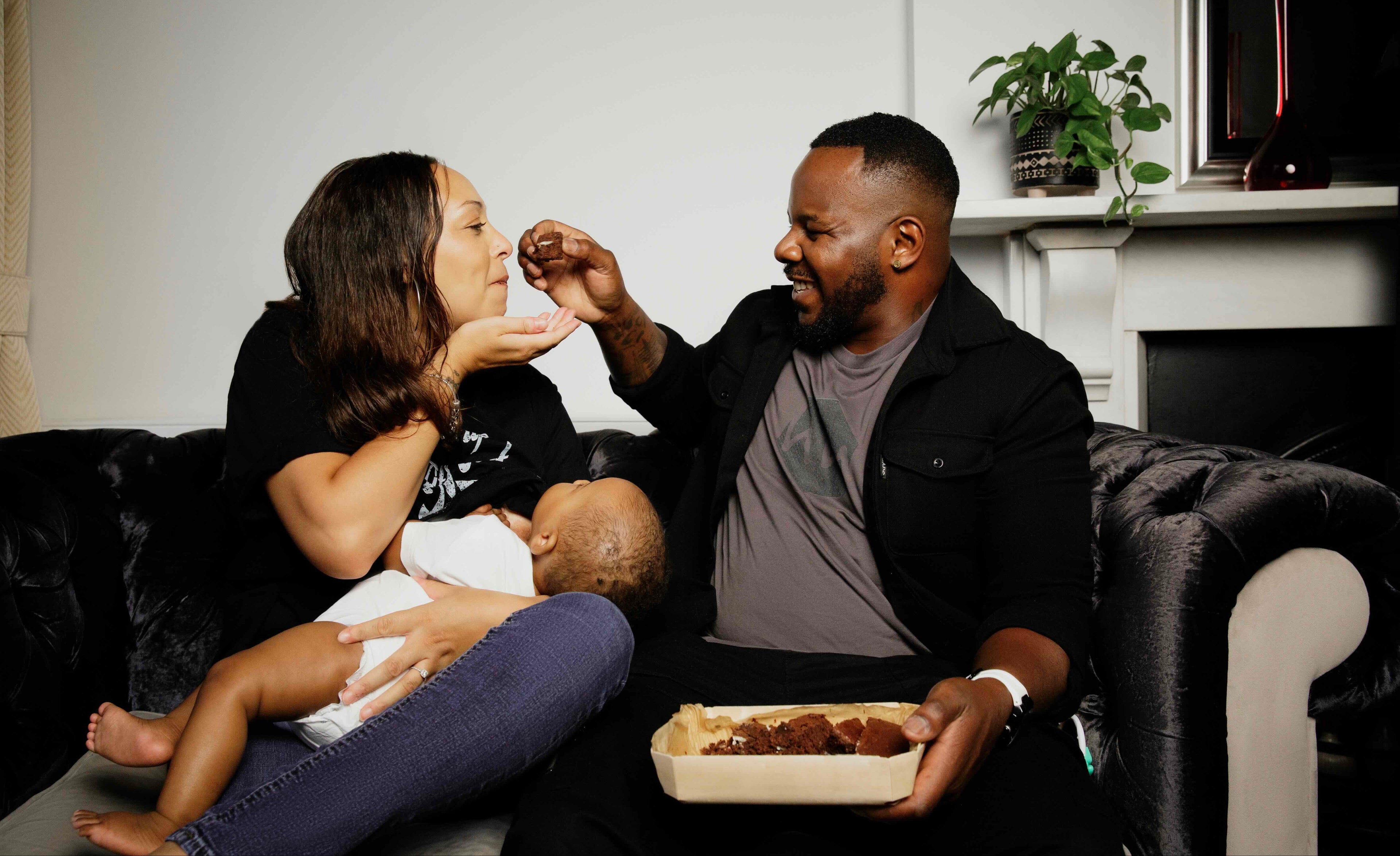 Family of three sitting on a couch with a woman breastfeeding a baby and a man feeding the mother chocolate brownies.