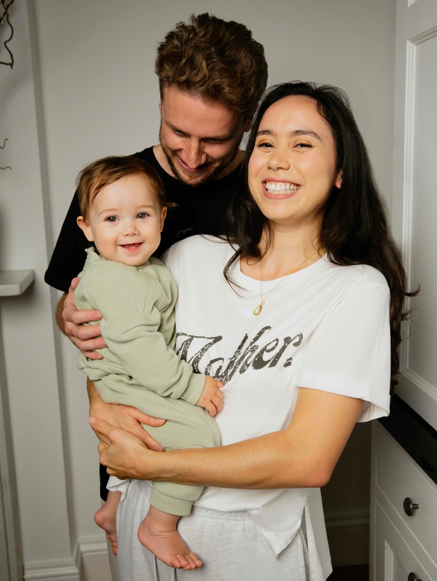 A mother, father and young toddler smiling at the camera. The mother is wearing a white human milk t-shirt with the word Mother on the front..