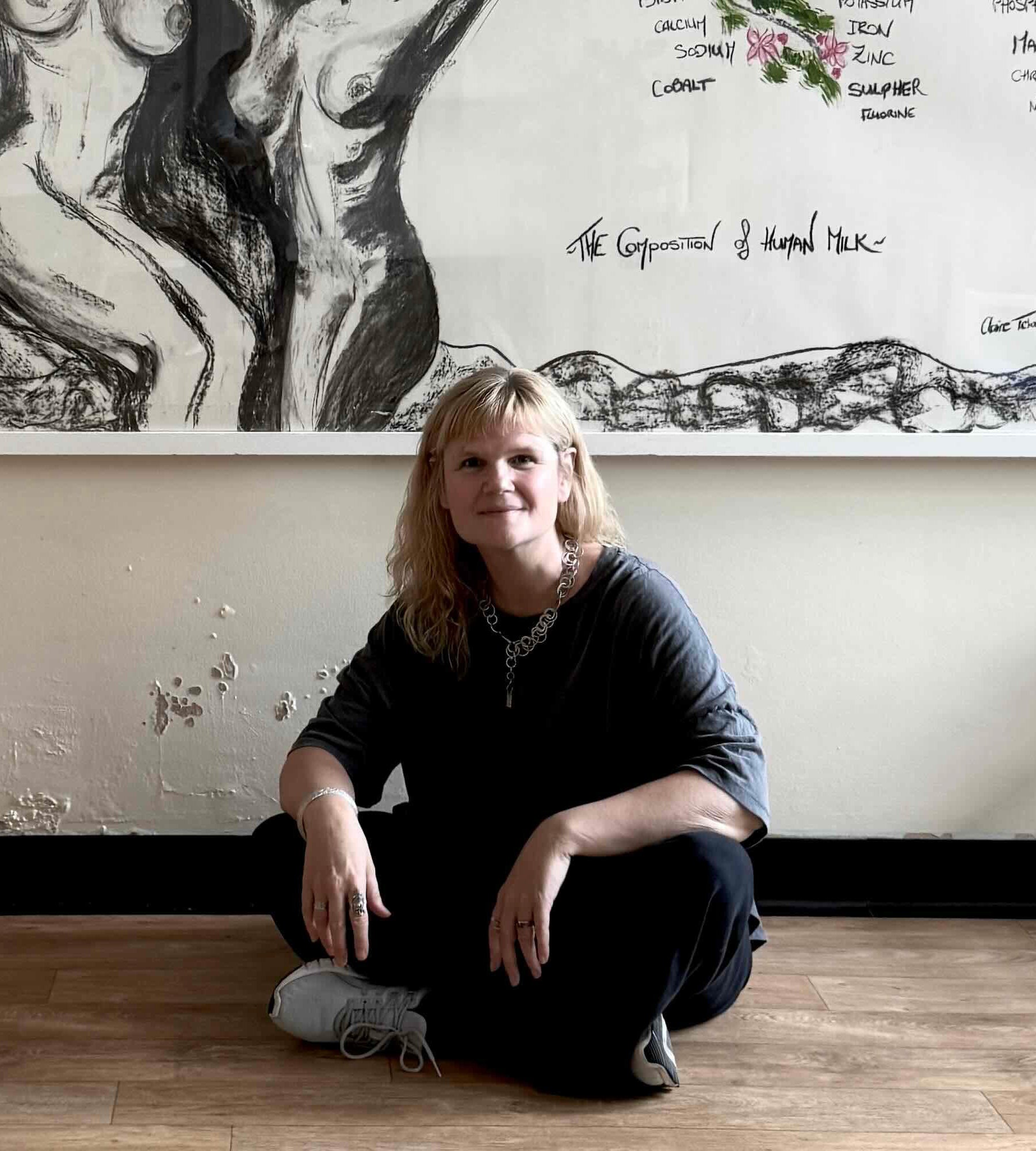 Claire Tchaikowski sitting on the floor in front of a large artwork on a wall representing the ingredients of breastmilk.