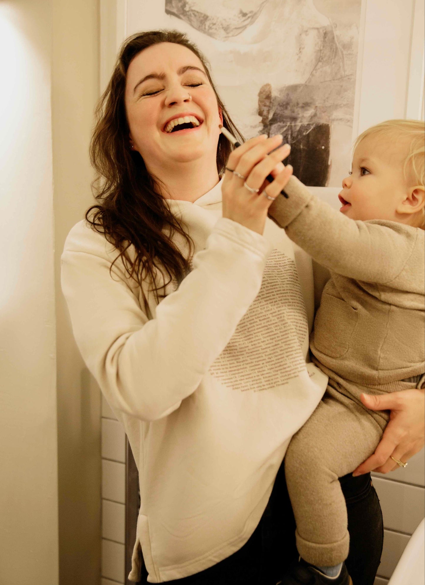 Woman wearing a Human Milk hoodie with the ingredients of breastmilk on it, laughing with a baby in a room with a painting on the wall.