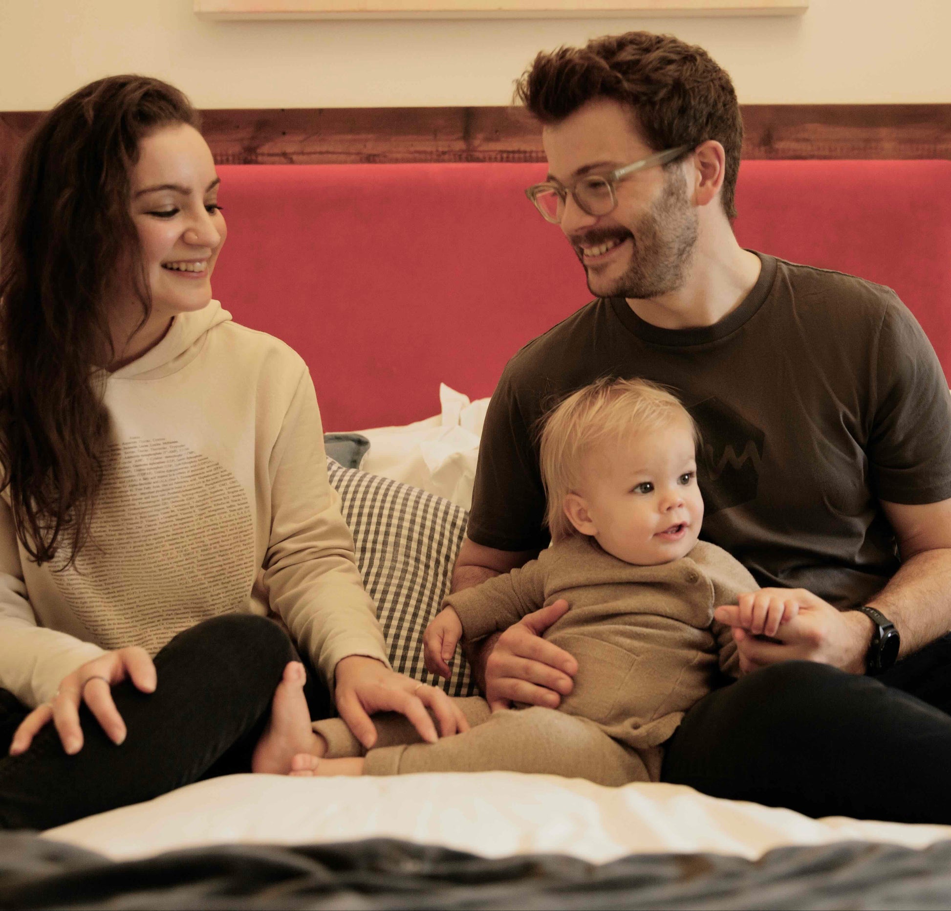 Mother, father and baby sitting on a bed laughing. Mother and father are wearing human milk tops.