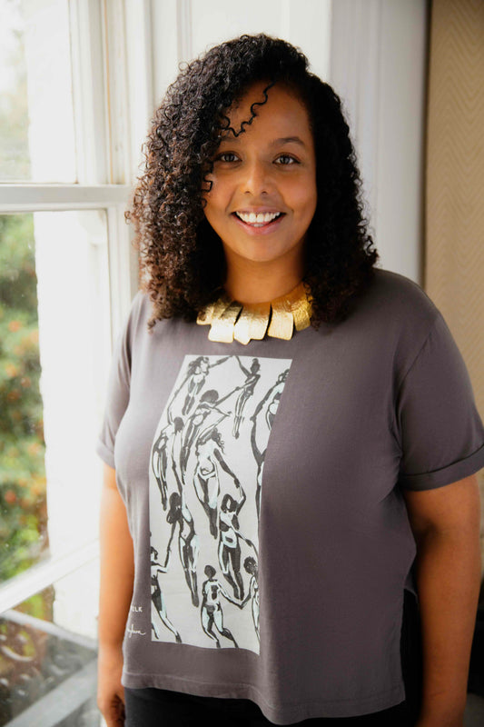 Woman wearing a grey human milk and Chloe Trayhurn t-shirt with graphic design, standing indoors near a window.