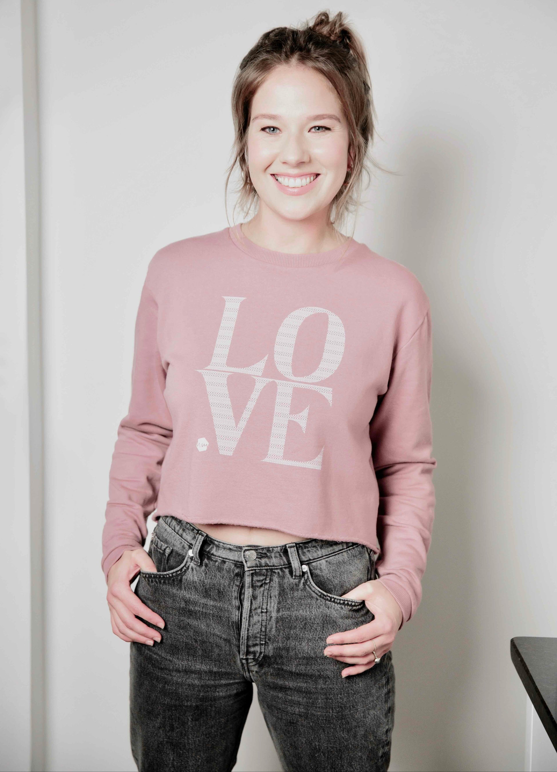 Woman wearing a pink 'LOVE' Human Milk sweatshirt and black jeans against a plain background
