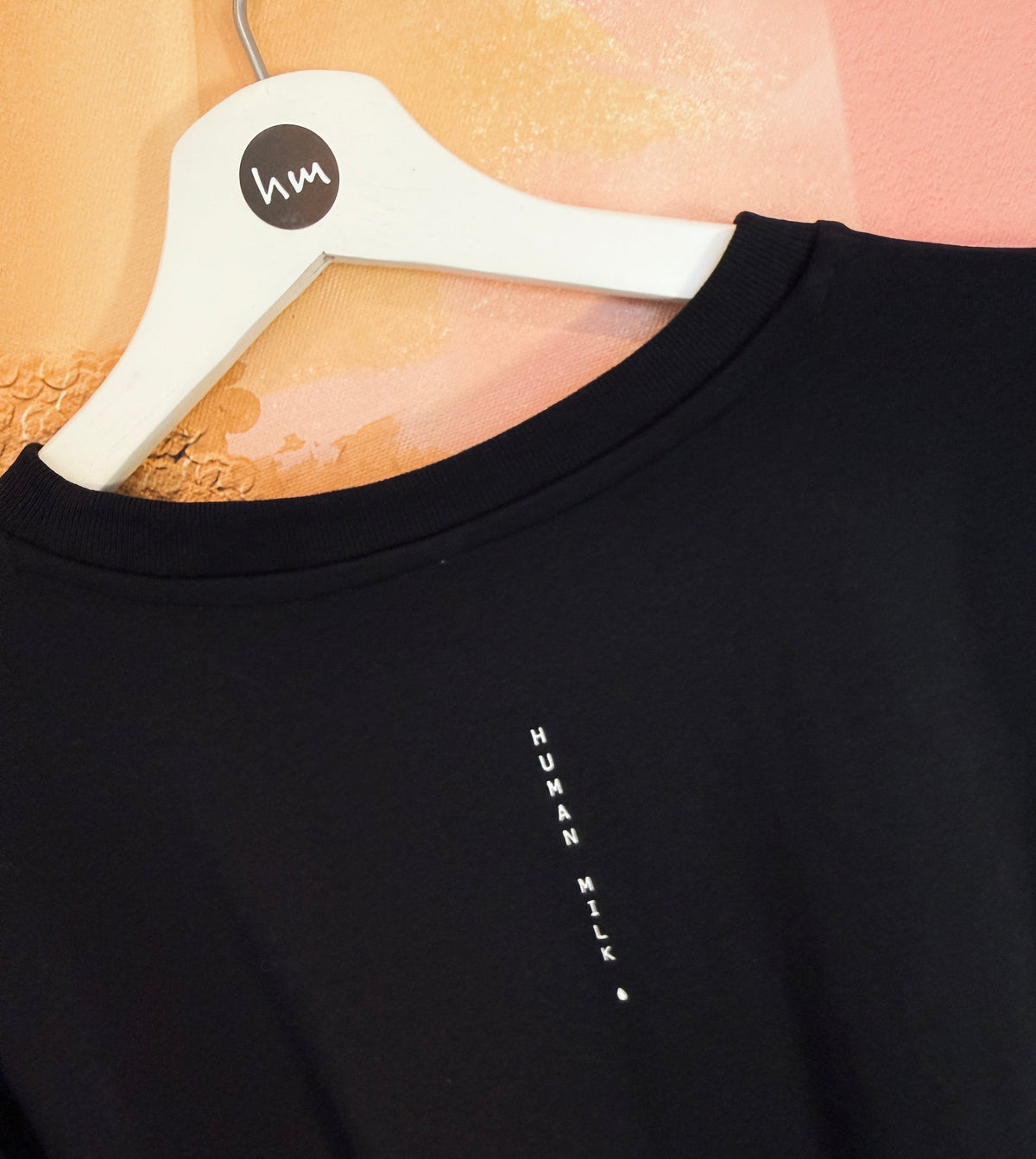 Black Human milk t-shirt with a small white logo on a hanger against a textured peach background