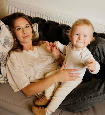 Woman wearing a human milk t-shirt sitting on a couch holding a baby, both wearing light-colored clothing.