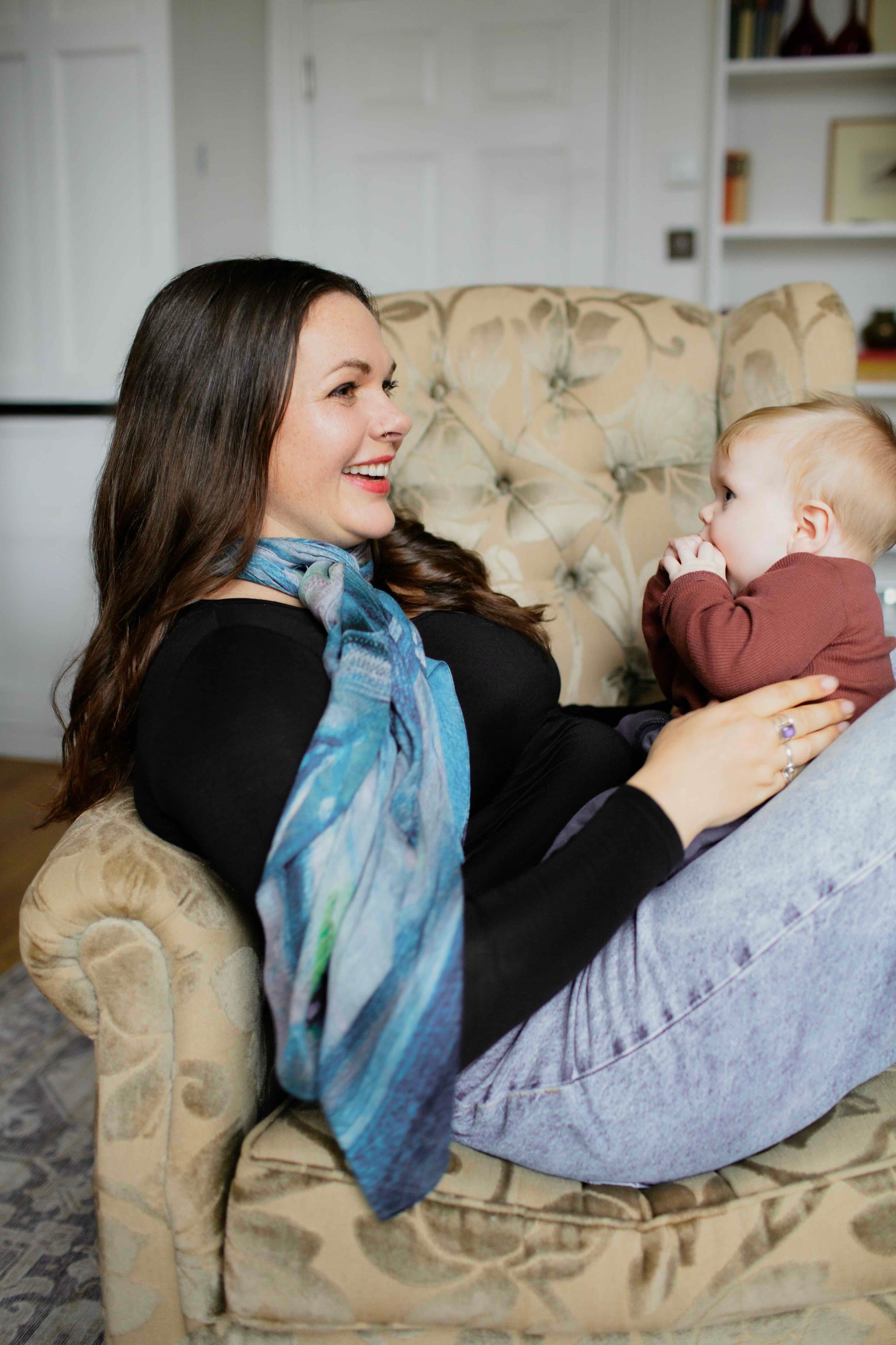 Woman sitting on a couch wearing a human milk scarf, holding a baby, both smiling