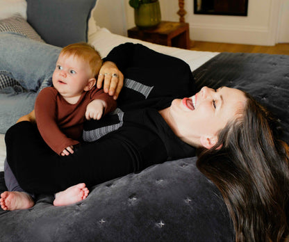 Woman wearing a Human Milk hoodie lying on a couch with a baby on her chest in a cozy living room.