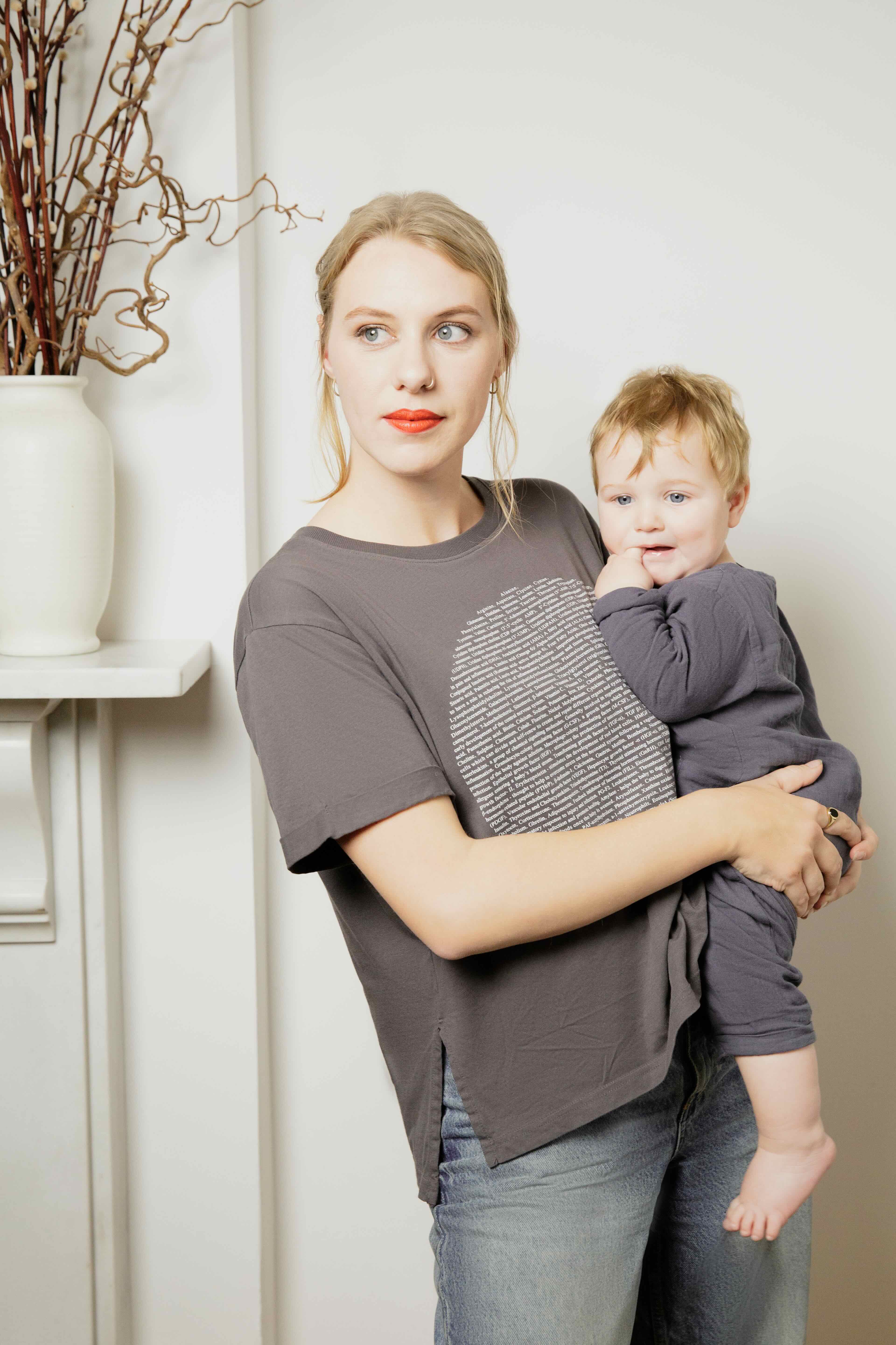 Woman wearing a human milk breastfeeding friendly t-shirt holding her child in a room with a white wall and decorative elements.