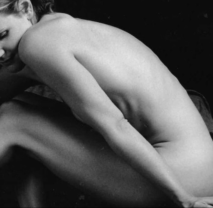 Black and white artistic photograph of a nude woman seated on a dark surface against a dark background. A Human Milk image.