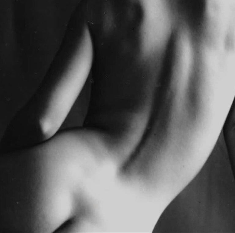 Black and white photograph of a nude woman seated on a surface against a dark studio backdrop, shown from the back and side. A Human Milk image.