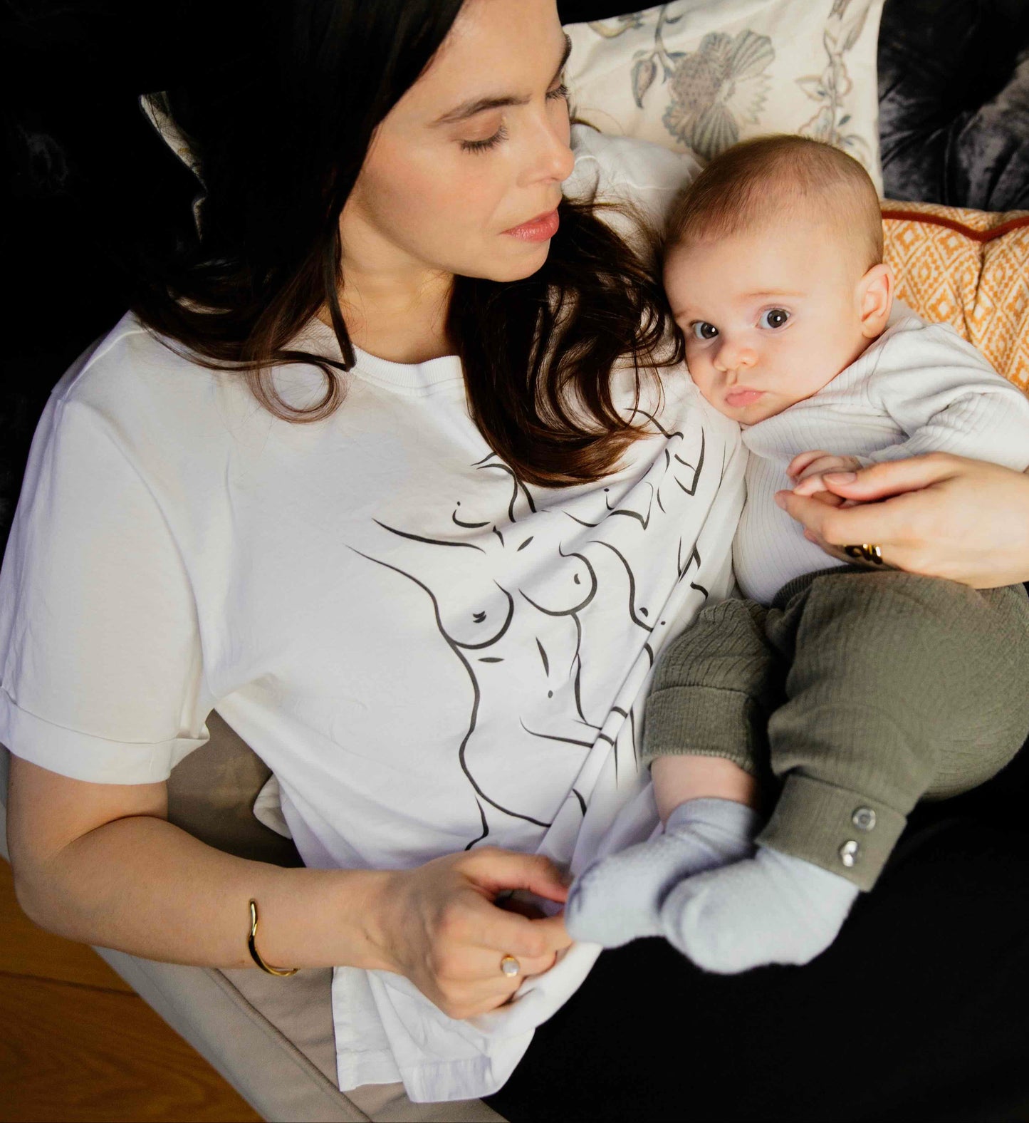 Woman wearing a Human Milk Woman T-shirt holding her baby on a couch in a cozy living room setting