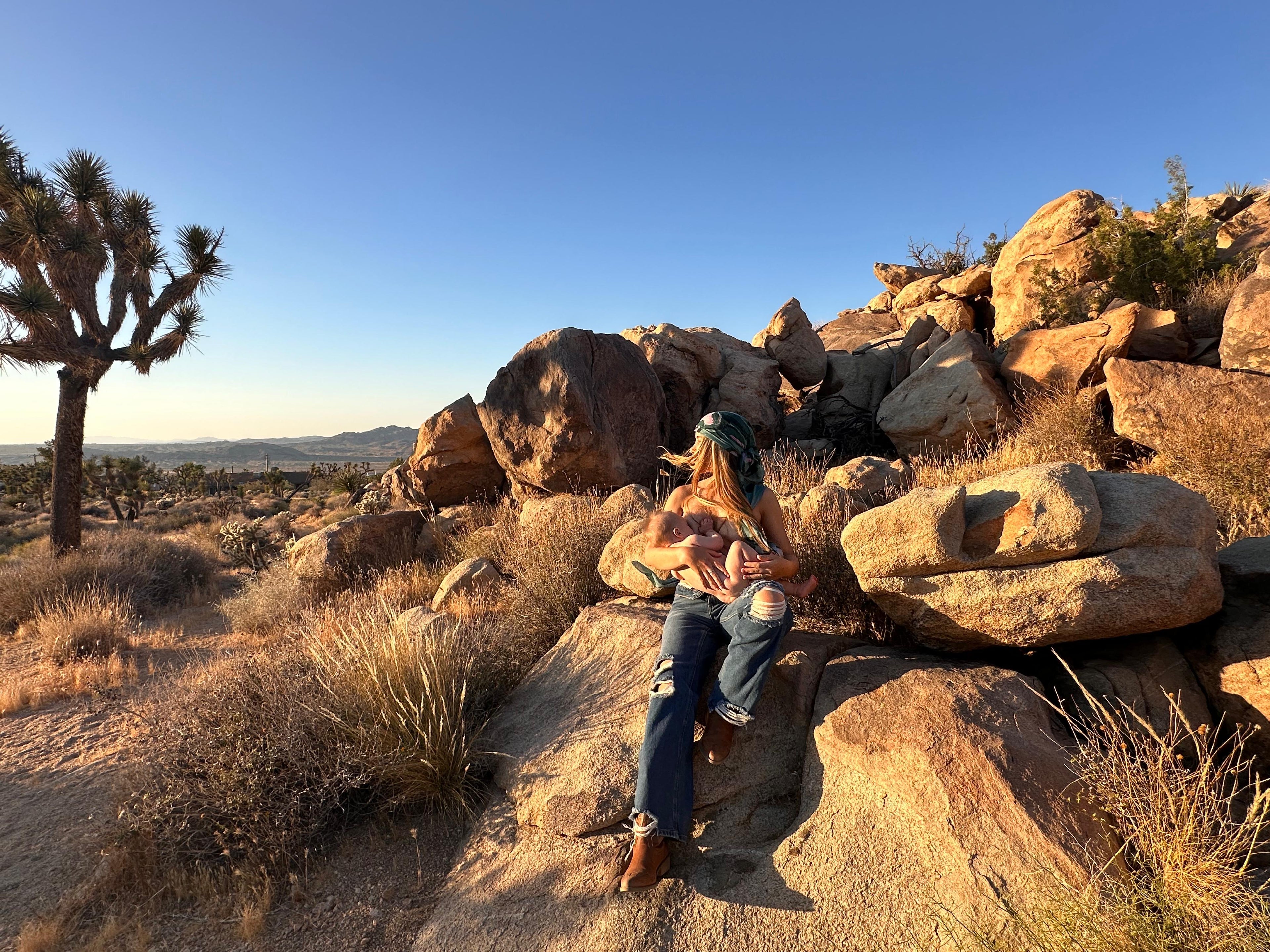 Mother sitting on a rock in a desert landscape, breastfeeding her baby, with Joshua trees and mountains in the background.