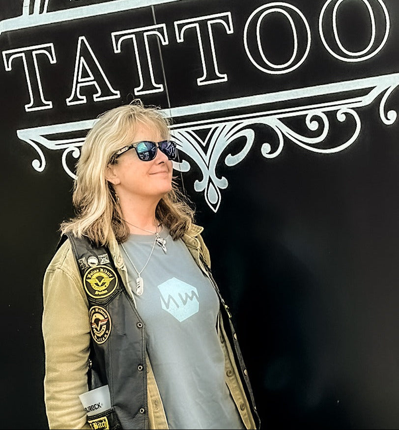 Lisa Ravenscroft wearing sunglasses and a jacket with patches in front of a 'Sideshow Tattoo' sign.